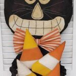 Primitive black cat doll holding candy corn pillows with stitched grin and striped bow tie.
