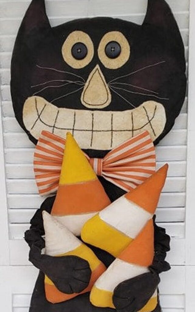 Primitive black cat doll holding candy corn pillows with stitched grin and striped bow tie.