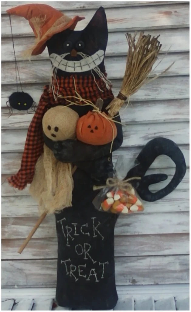 Primitive Halloween black cat doll dressed as a witch, holding a broom, pumpkin, ghost and candy corn bag, with stitched ‘Trick or Treat’ on the body.