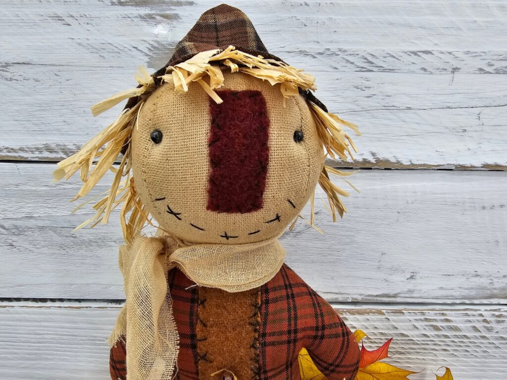 Close‑up of primitive folk art scarecrow showing stitched facial features, black bead eyes, and wool nose