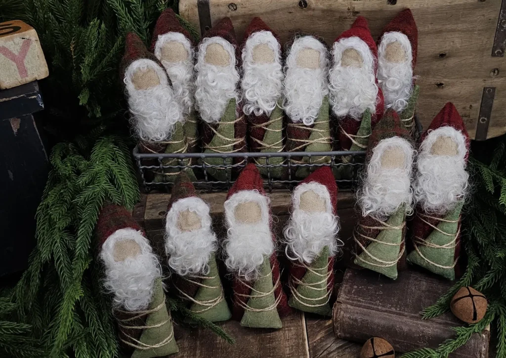 Primitive Santa tuck in red wool sack holding hand‑painted tree, with needle‑felted wool beard and simple folk‑art Christmas styling.