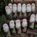 Primitive Santa tuck in red wool sack holding hand‑painted tree, with needle‑felted wool beard and simple folk‑art Christmas styling.
