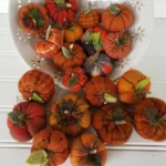 Mini flannel woolies pumpkins spilling out of a bowl, showing assorted autumn fabrics, wood stems, and wool leaves.