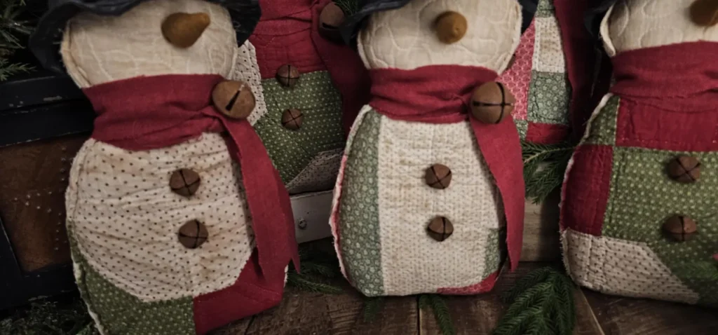 Grouping of primitive quilted snowmen displaying rag‑stuffed bodies made from vintage patchwork quilts, wool scarves with rusty jingle bells, and rustic farmhouse details.