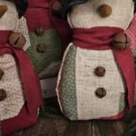 Grouping of primitive quilted snowmen displaying rag‑stuffed bodies made from vintage patchwork quilts, wool scarves with rusty jingle bells, and rustic farmhouse details.