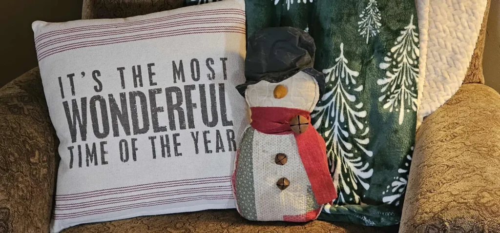 Primitive quilted snowman displayed on a cozy armchair with holiday pillow and green winter blanket, showing how the snowman can be used in farmhouse Christmas décor.