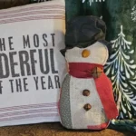Primitive quilted snowman displayed on a cozy armchair with holiday pillow and green winter blanket, showing how the snowman can be used in farmhouse Christmas décor.